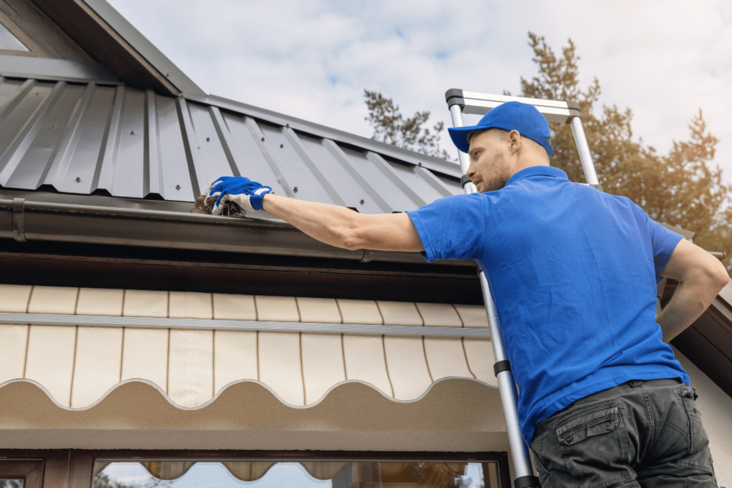 A man in a blue shirt and hat, standing on a ladder, wearing a glove to clean out a home gutter