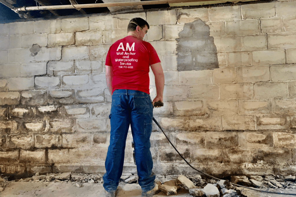 Man wearing a red AM Wall Anchor & Waterproofing shirt is drilling in a basement